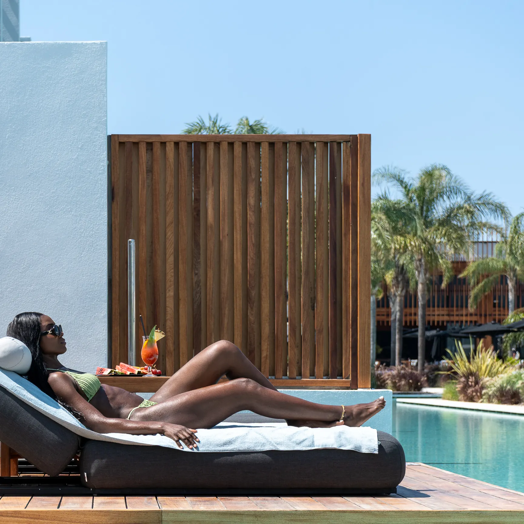 Woman in sunglasses reclining on a poolside daybed beside a slatted timber screen and palm trees
