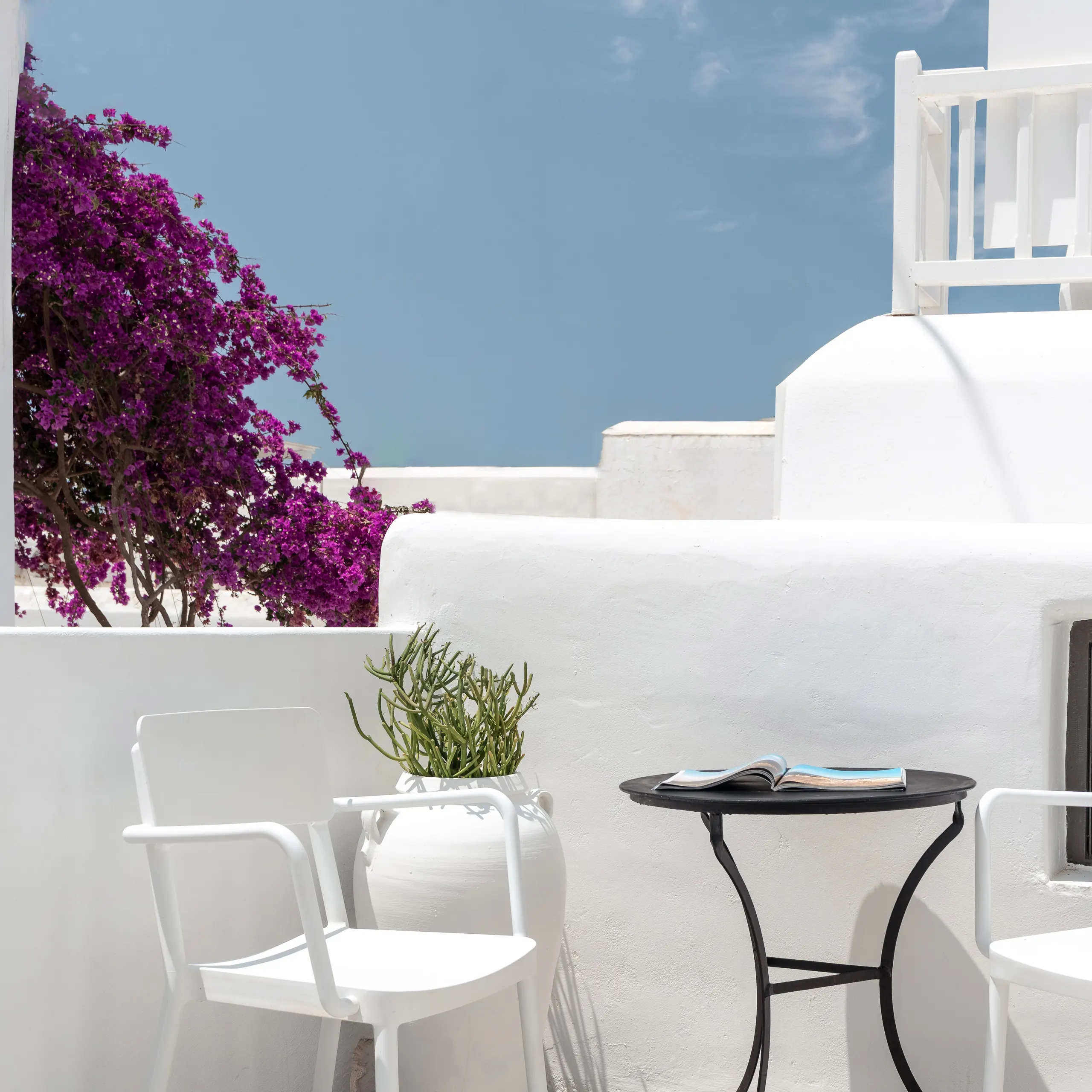 White Cycladic terrace with bougainvillea, white chairs and a black bistro table under a blue sky