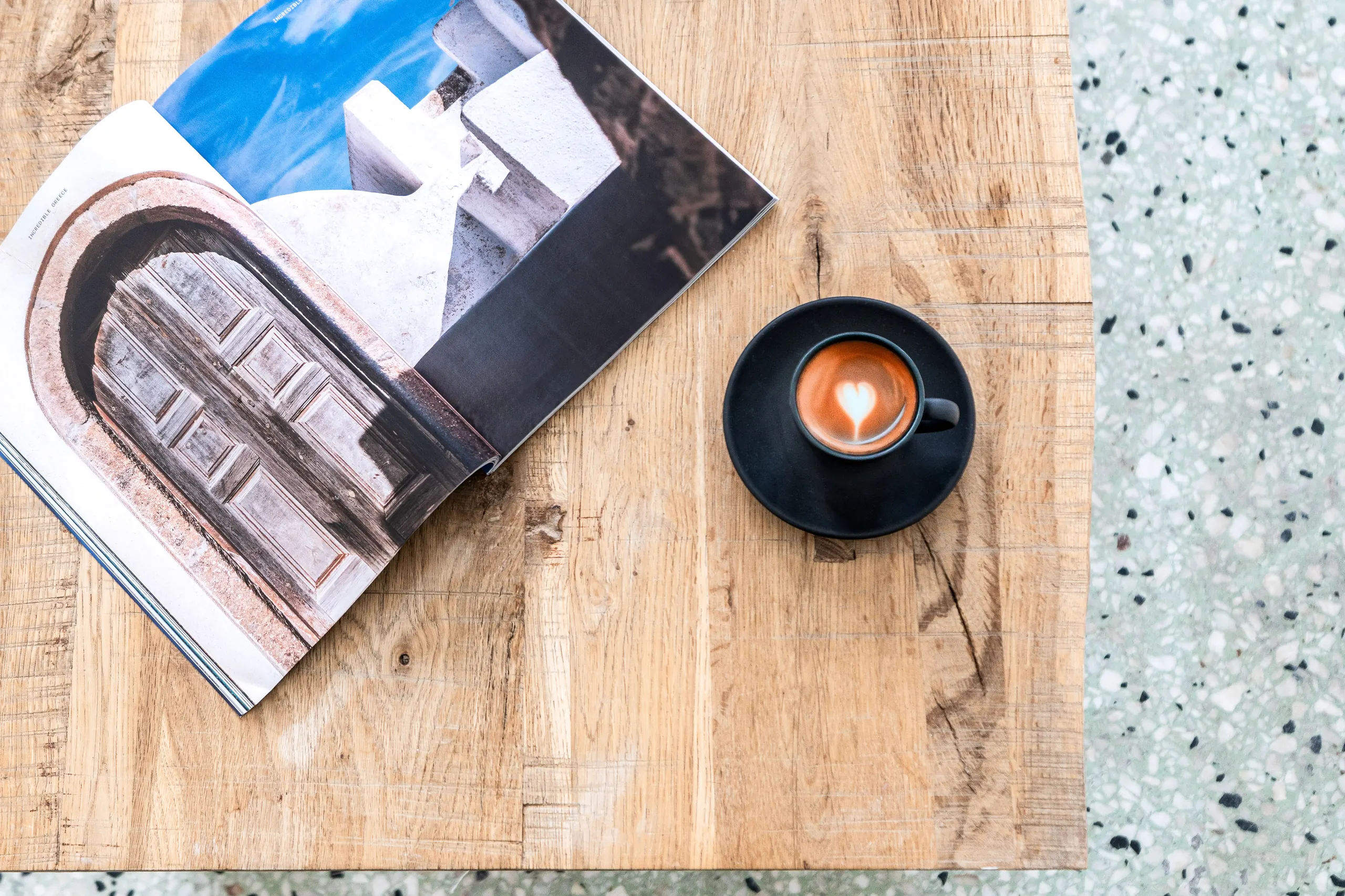 Top-down view of an espresso with latte art beside an open travel magazine on an oak table