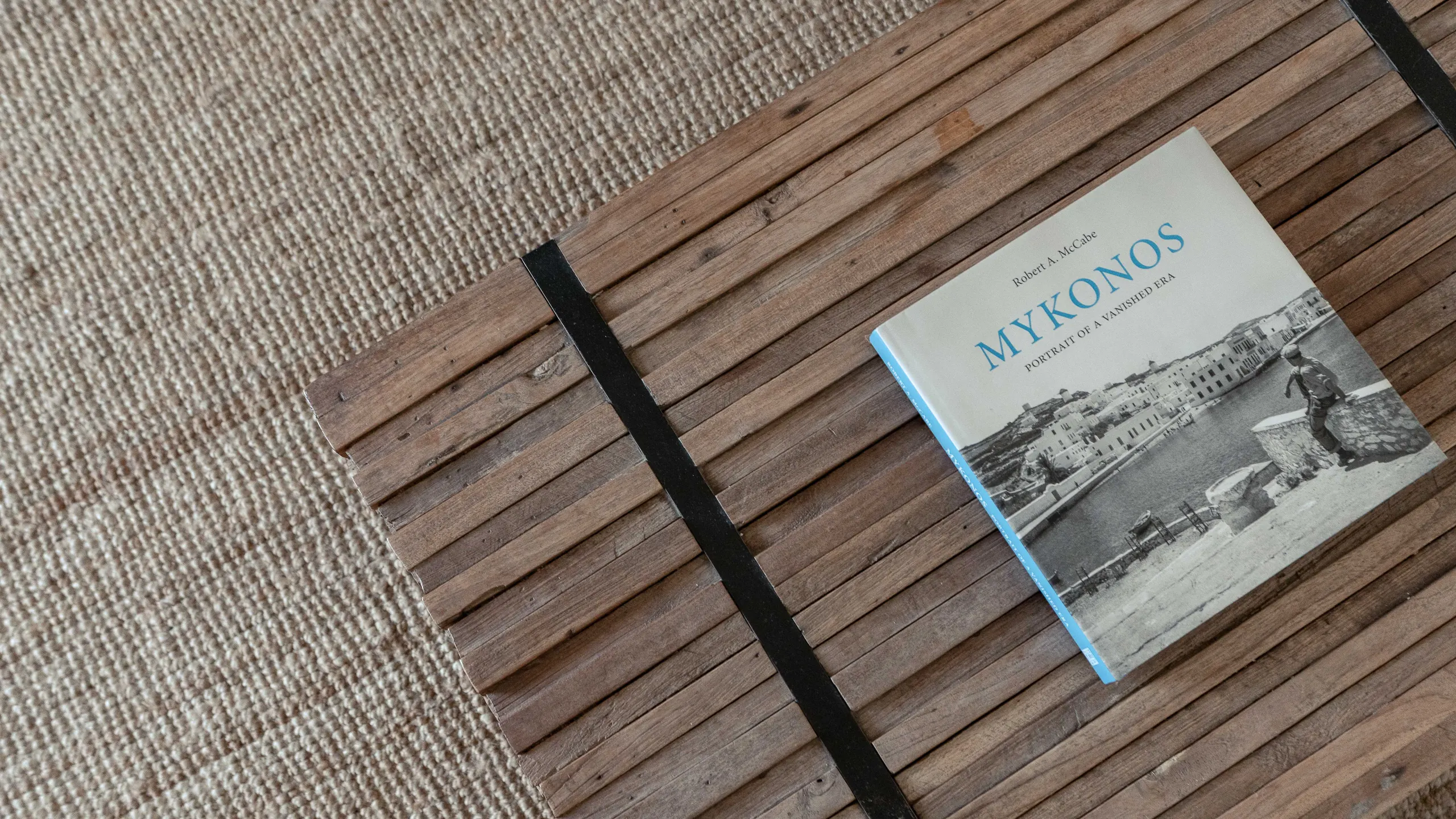 A book titled Mykonos, Portrait of a Vanished Era, resting on a slatted wooden table over a jute rug