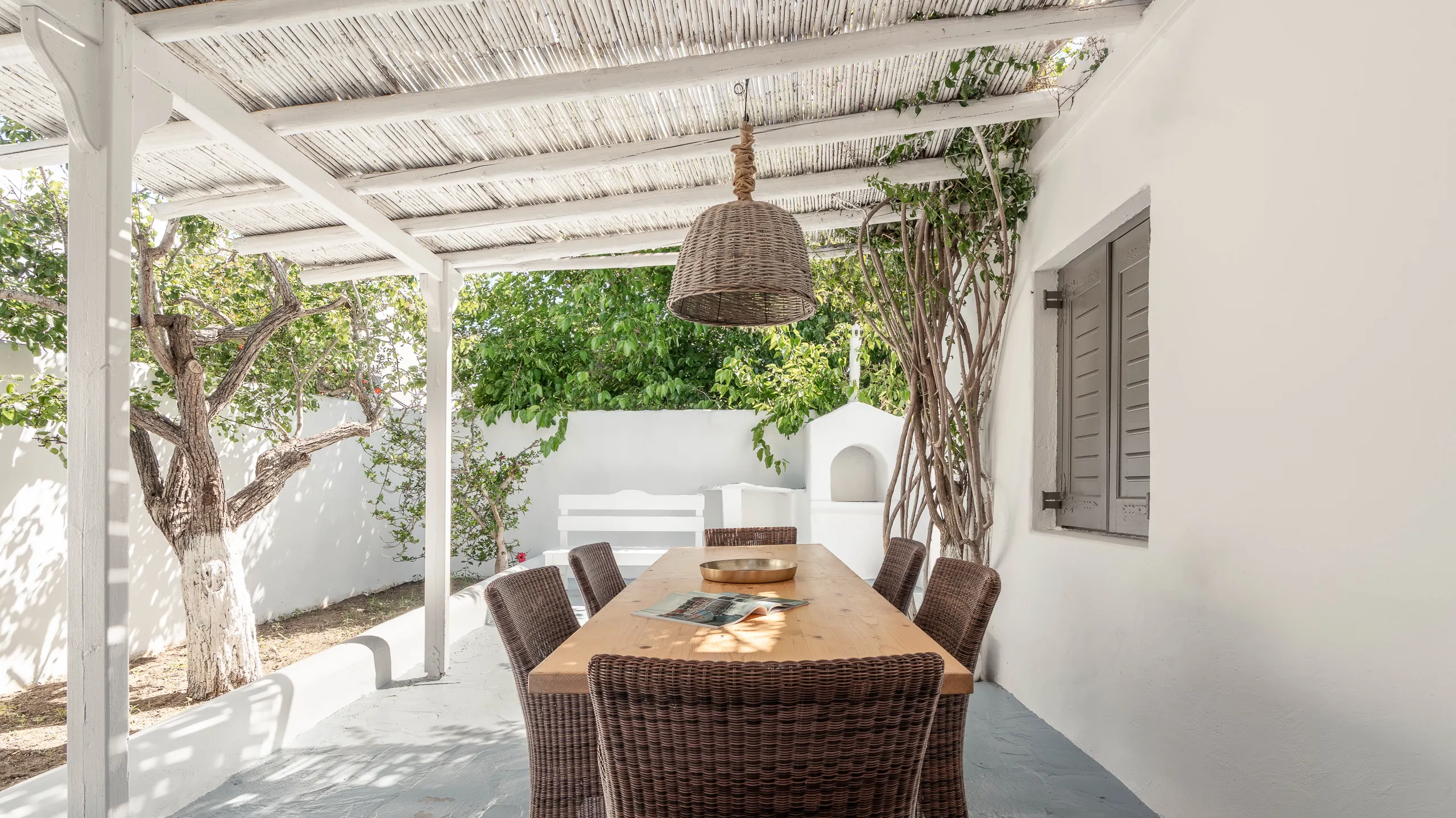 Shaded dining terrace with wicker chairs, a long wooden table and a woven pendant lamp under a reed pergola