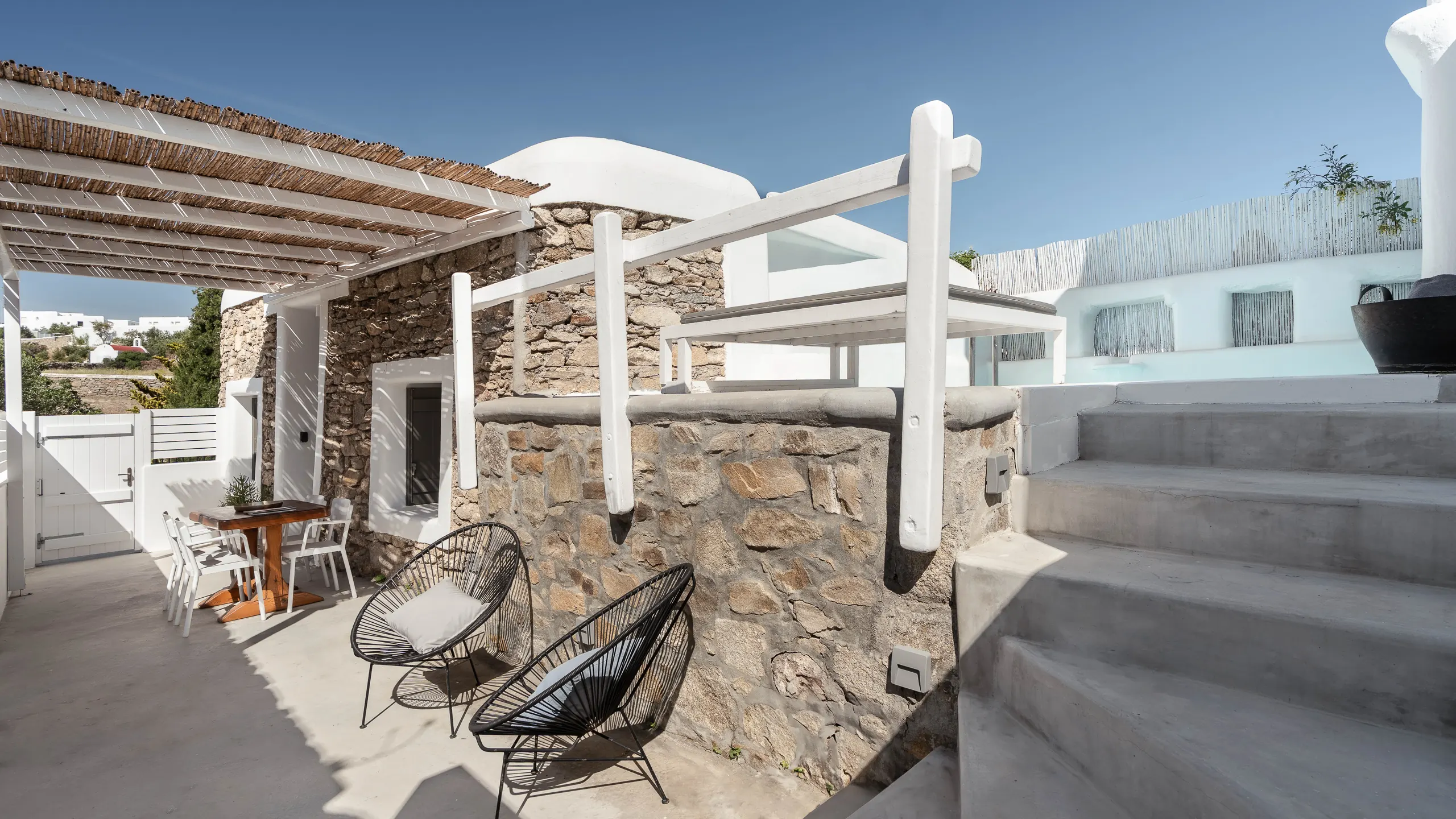Cycladic courtyard with stone walls, a white pergola, wire lounge chairs and concrete steps